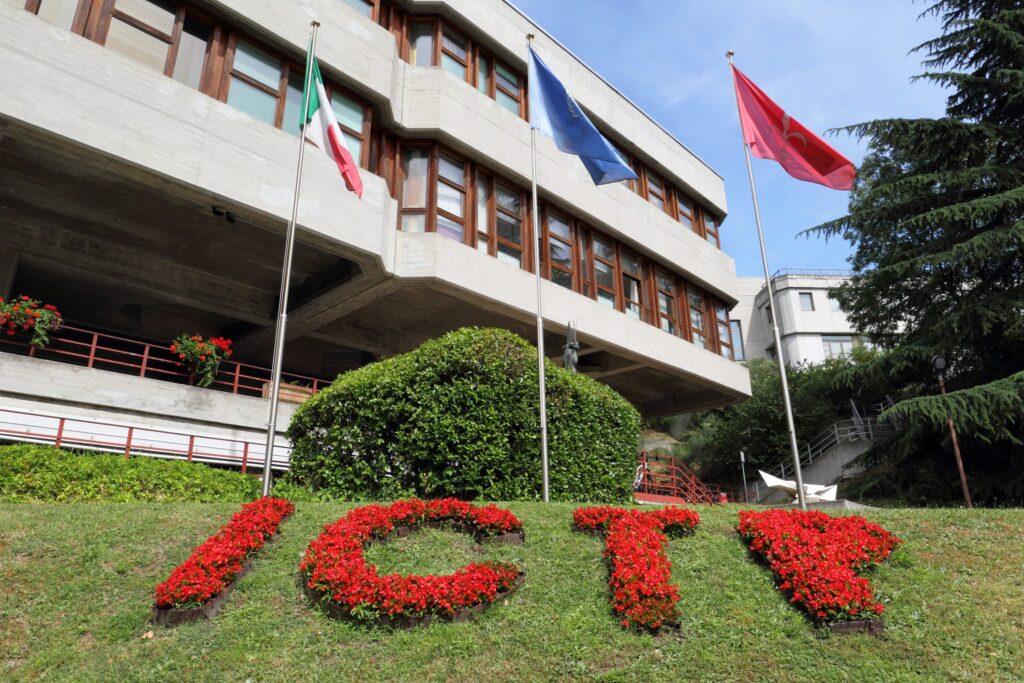 The front facade of the main building named Leonardo, with a floral presentation of ICTP. The three flags in the backdrop are (from L to R) those of Italy, UNESCO and Trieste.